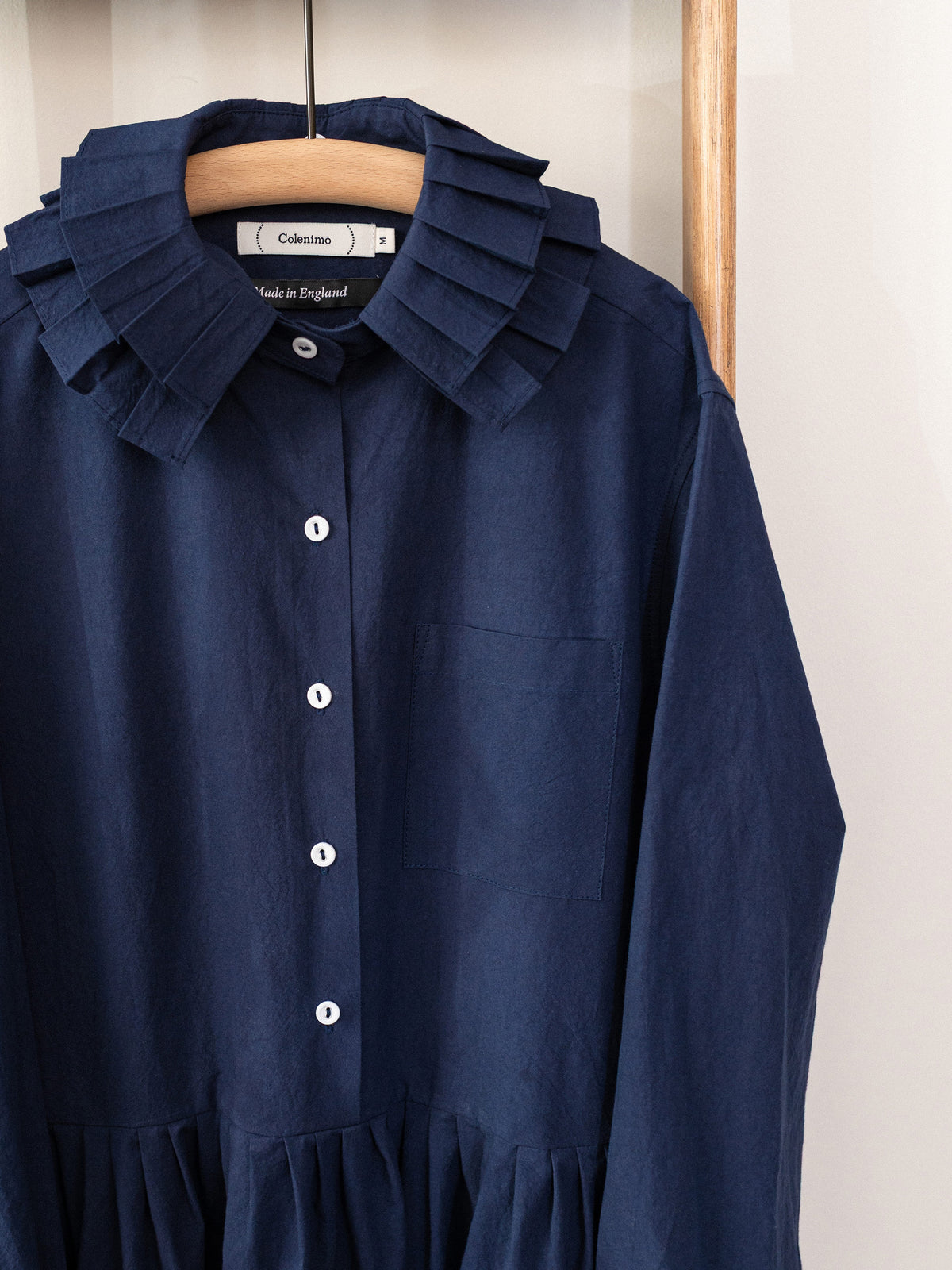 A navy blue long-sleeve shirt dress with a ruffled collar and white buttons, hanging on a wooden hanger against a light wall.