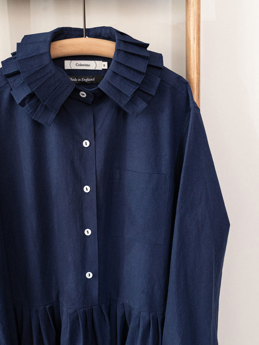 A navy blue long-sleeve shirt dress with a ruffled collar and white buttons, hanging on a wooden hanger against a light wall.