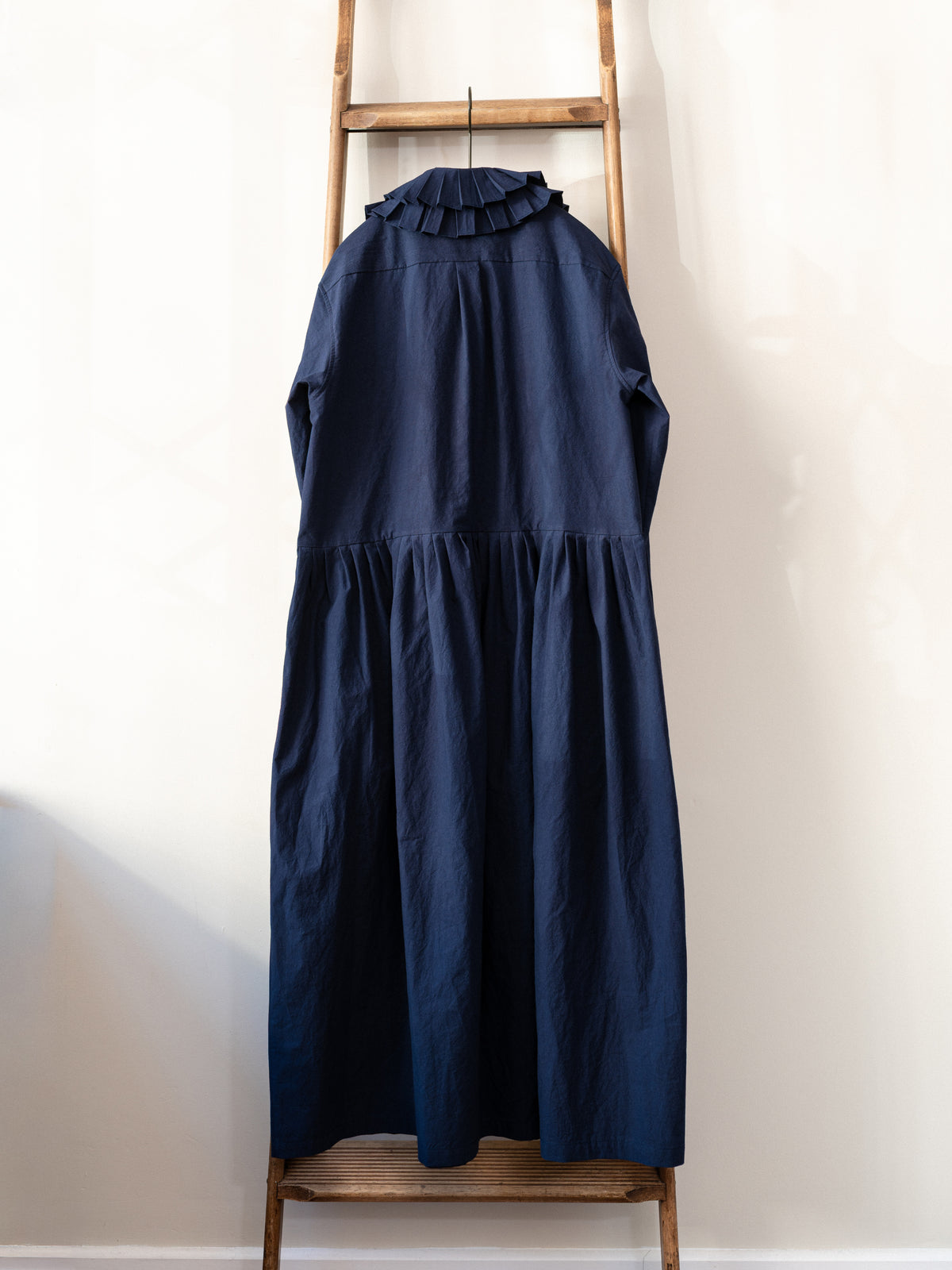 A dark navy blue tiered dress with a ruffled mock neck collar, hanging on a wooden ladder against a plain off-white wall.