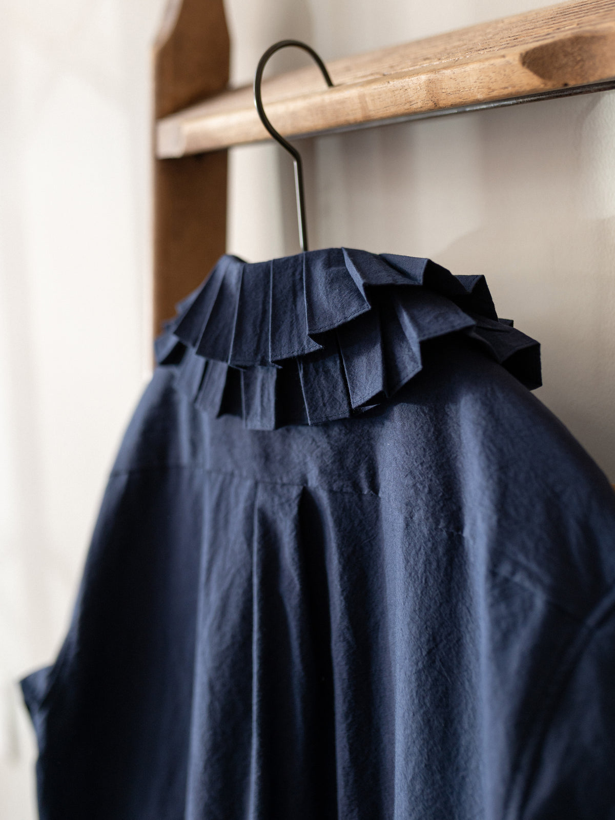 A navy blue blouse with a dramatic layered ruffle collar, hanging on a black wire hanger from a wooden clothes rack.