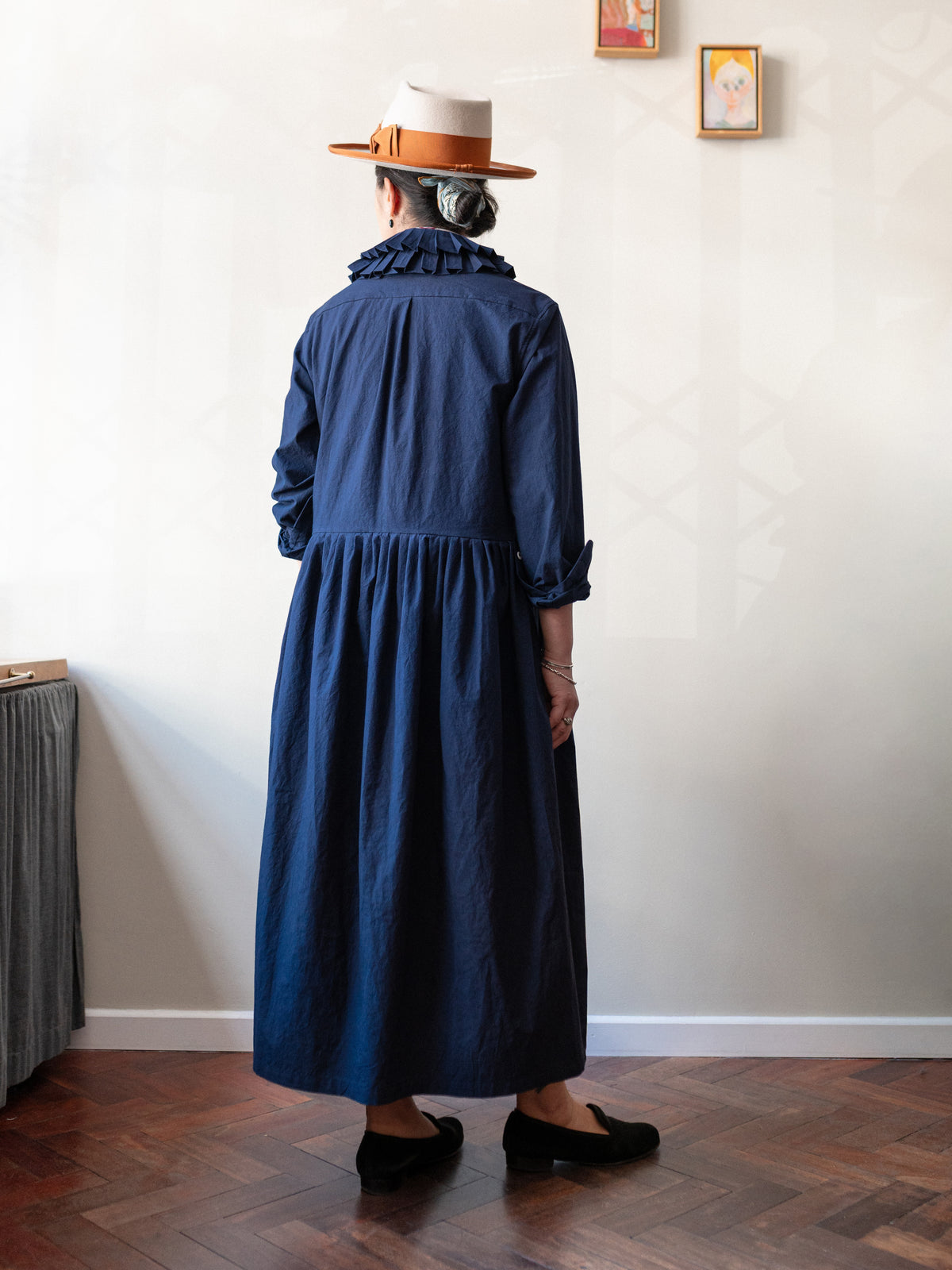 A person standing with their back to the camera wearing a long navy blue dress, a white and orange hat, and black shoes in an indoor room with a wooden floor and white wall.