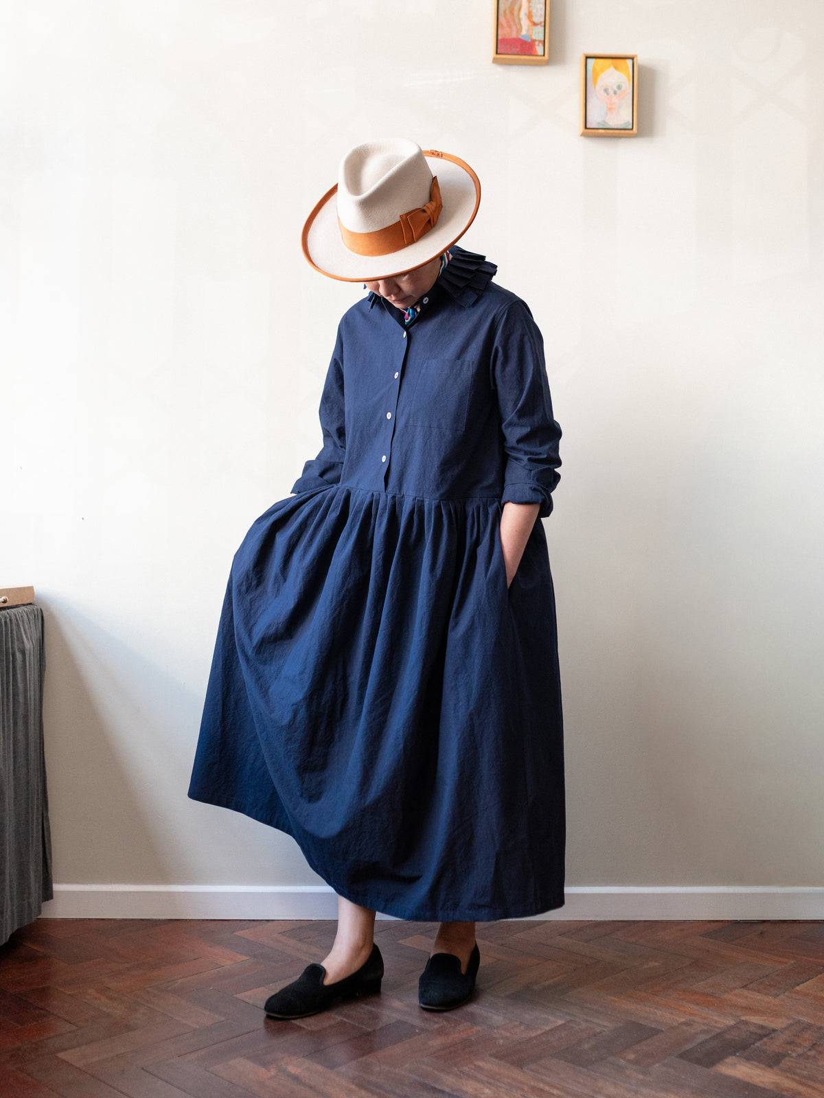 A person wearing a dark navy blue hooded midi dress with a gathered waist, paired with a cream fedora hat with a brown band and black slip-on shoes, standing in a room with a light-colored wall and wooden parquet floor.