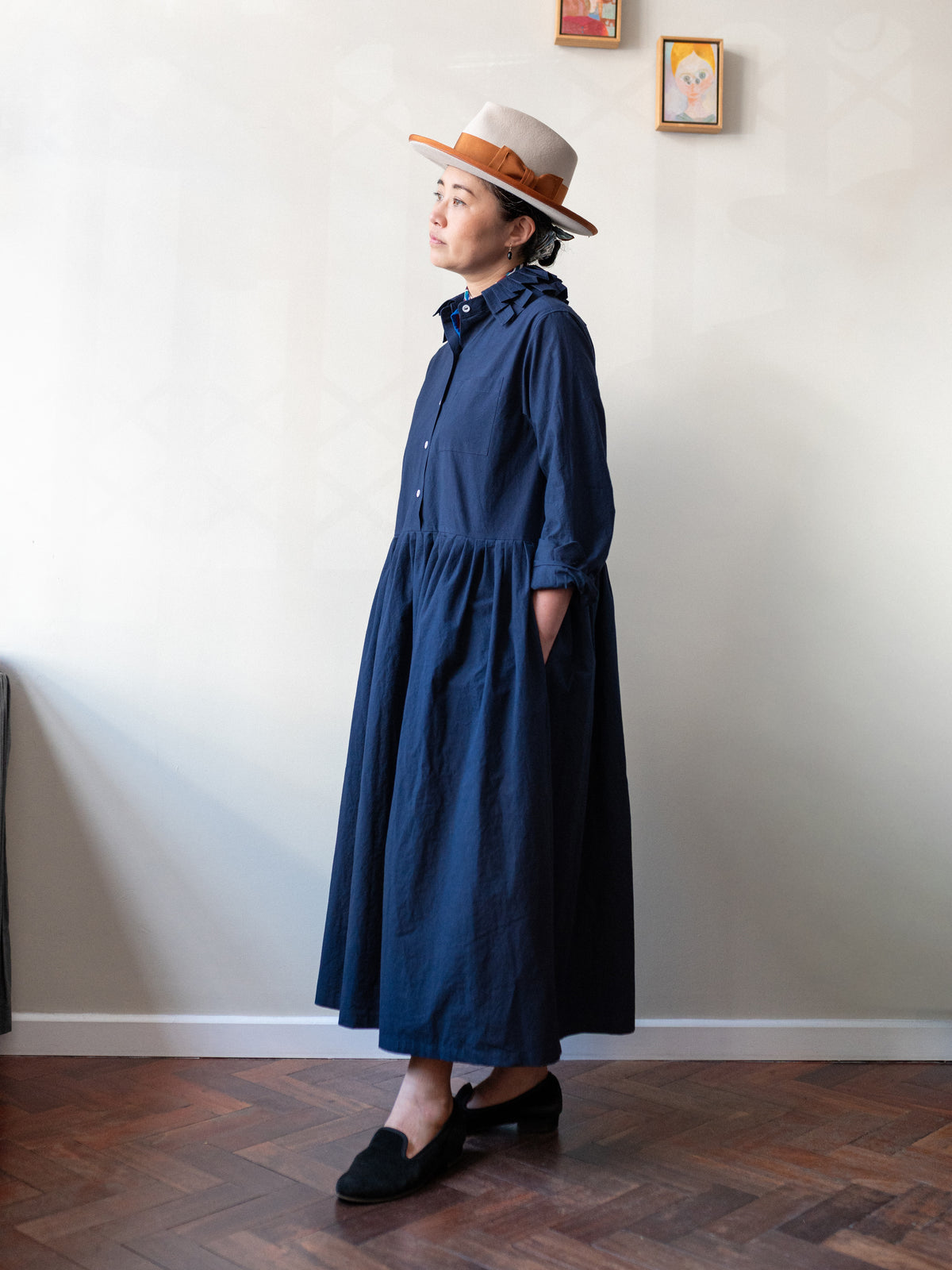 A woman wearing a navy blue long-sleeve midi dress with a gathered waist, paired with a cream-colored straw hat with a brown band and black slip-on shoes, standing in a room with a light-colored wall and wooden parquet floor.