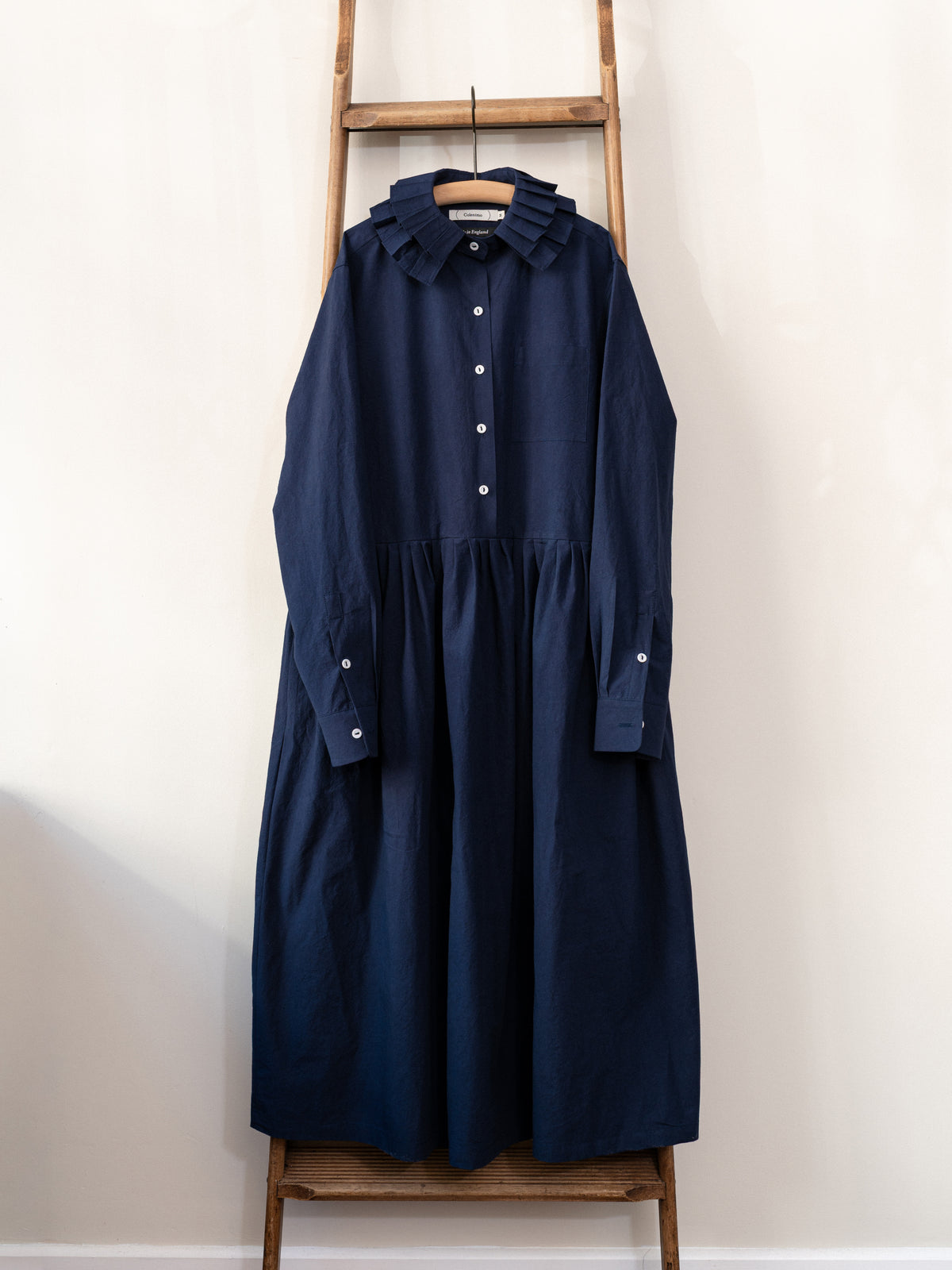 A dark navy blue long-sleeve dress with a ruffled collar and button front, hanging on a wooden hanger from a wooden ladder against a plain off-white wall.