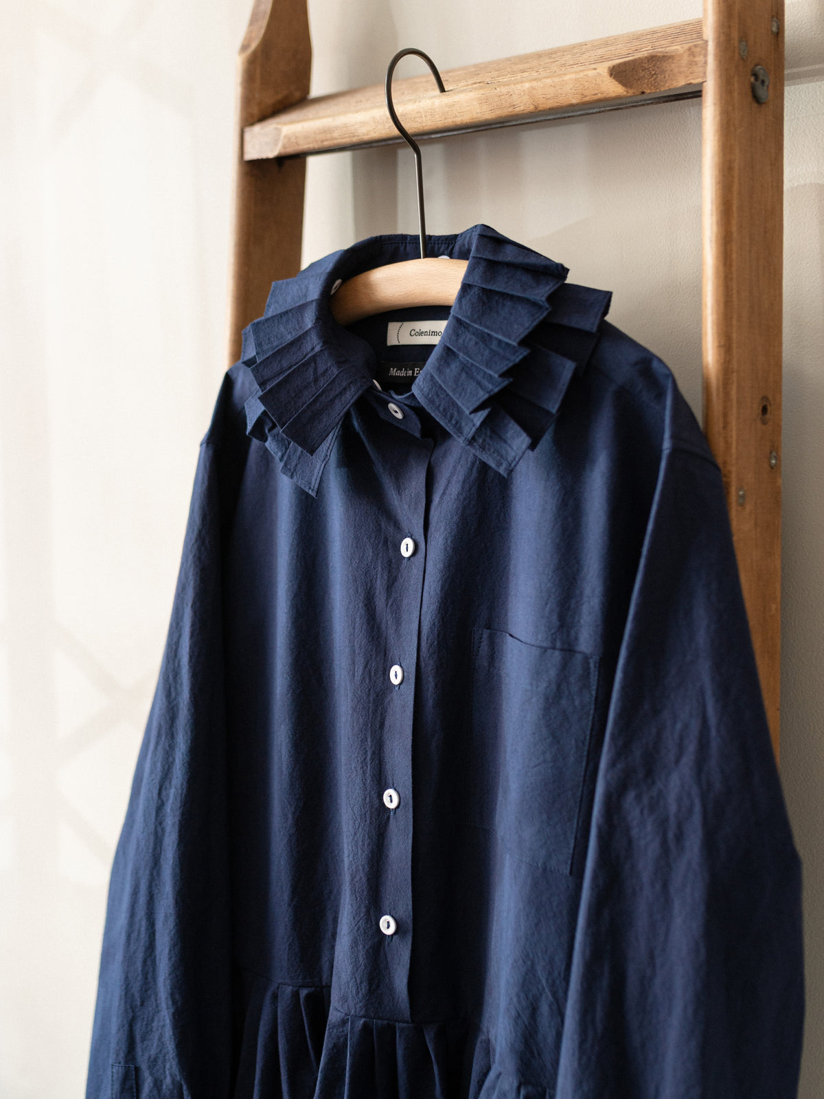 A navy blue long-sleeve button-up dress with a unique layered ruffle collar, hanging on a wooden hanger from a rustic wooden clothes rack.
