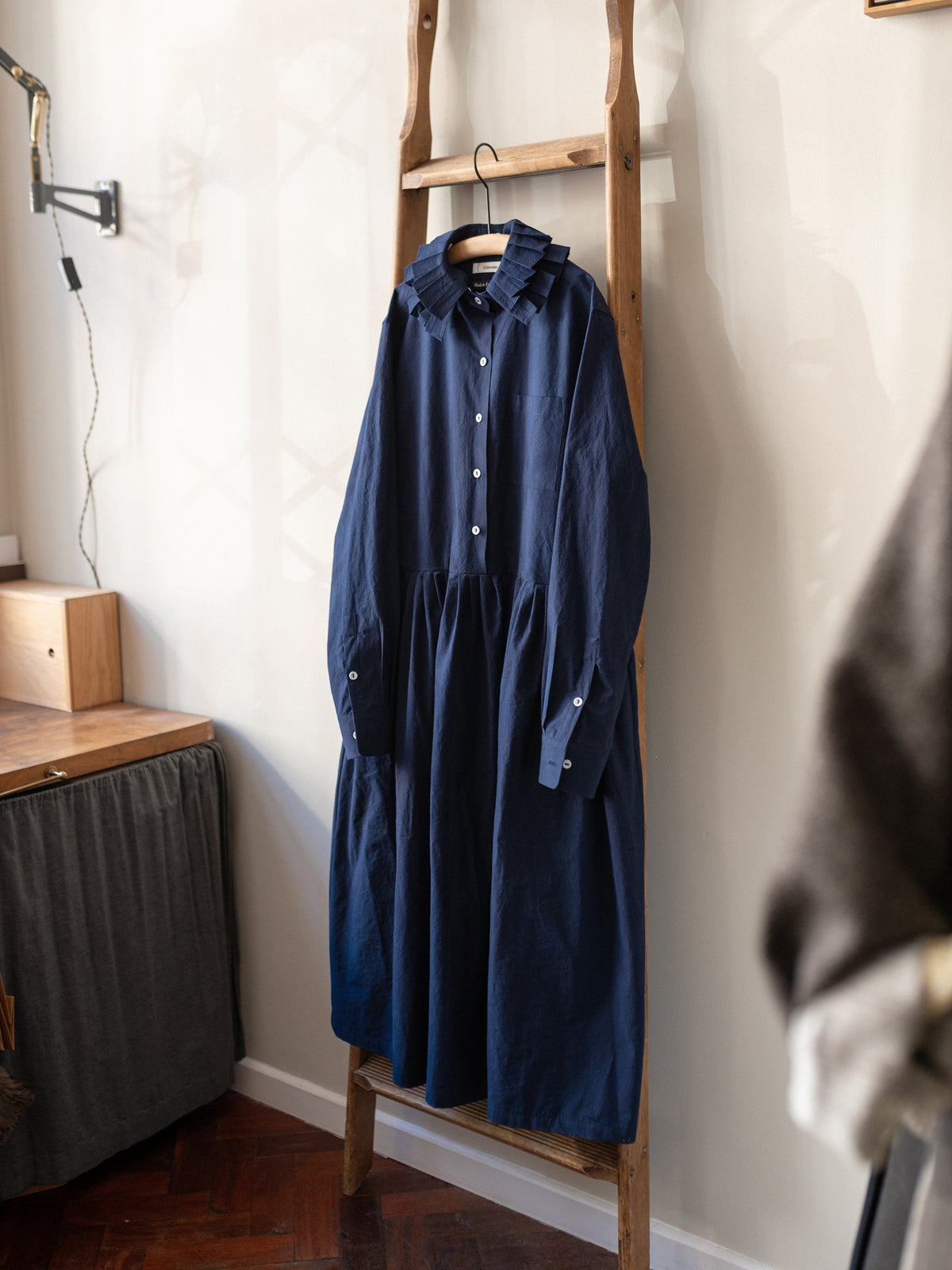 A dark navy blue long-sleeve shirt dress with a ruffled collar hangs on a wooden ladder rack in a home interior setting.