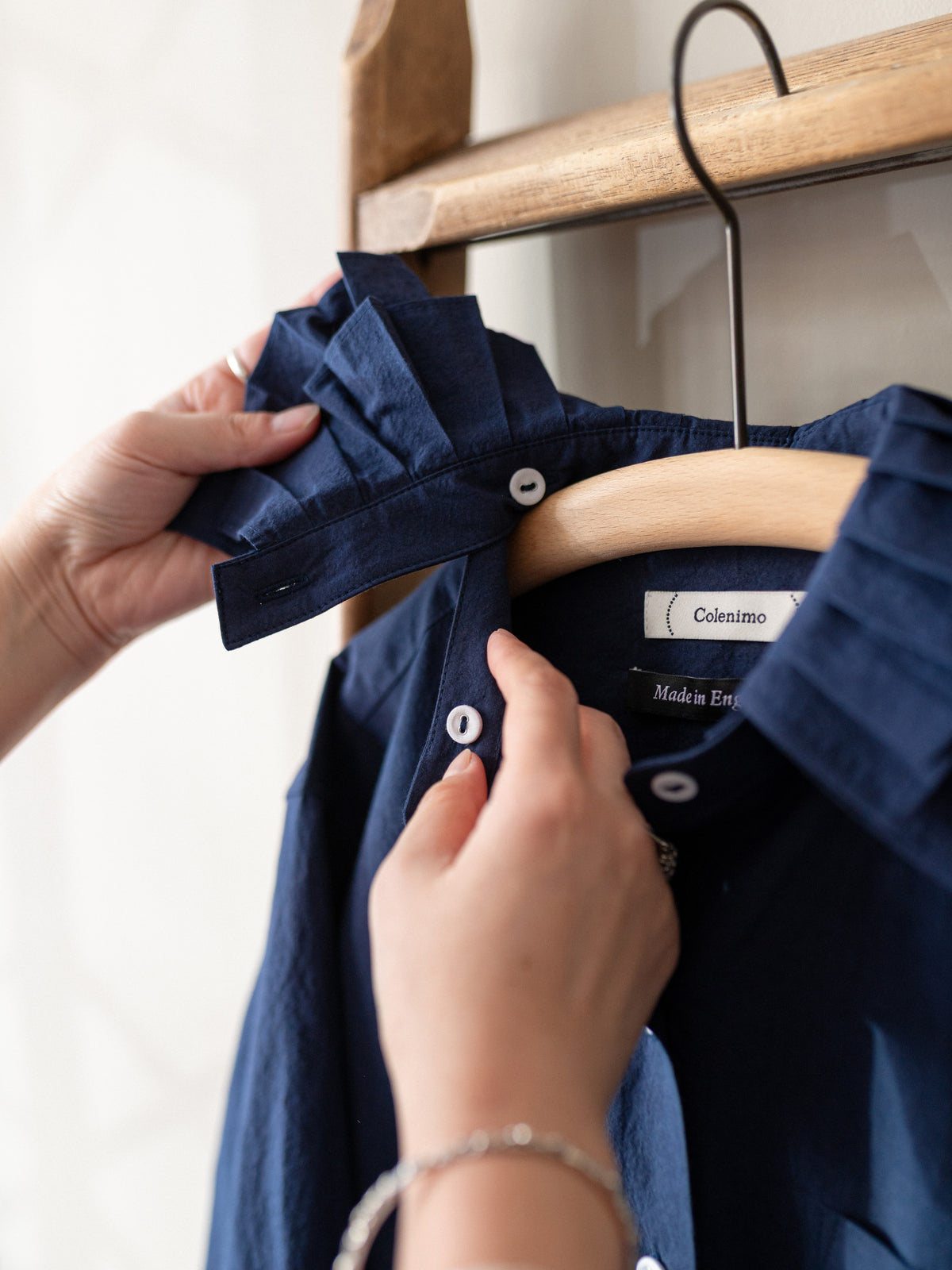 A close-up of a navy blue button-down shirt with a pleated collar detail, hanging on a wooden hanger. Hands are shown holding the collar and adjusting the shirt, with a wooden clothing rack visible in the background.